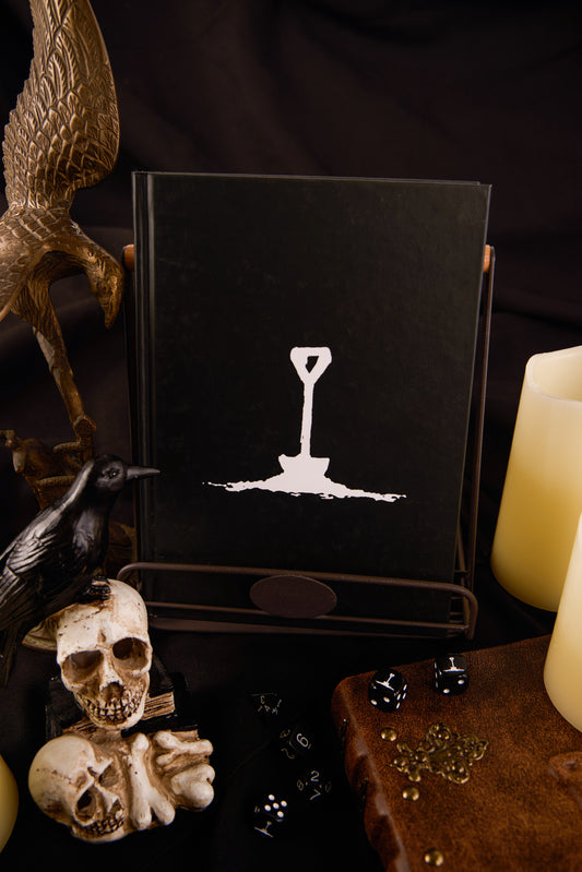 Best Left Buried Deeper cover product photography showing a black cover with white spade logo. Surrounded by dice, candles and other spooky props